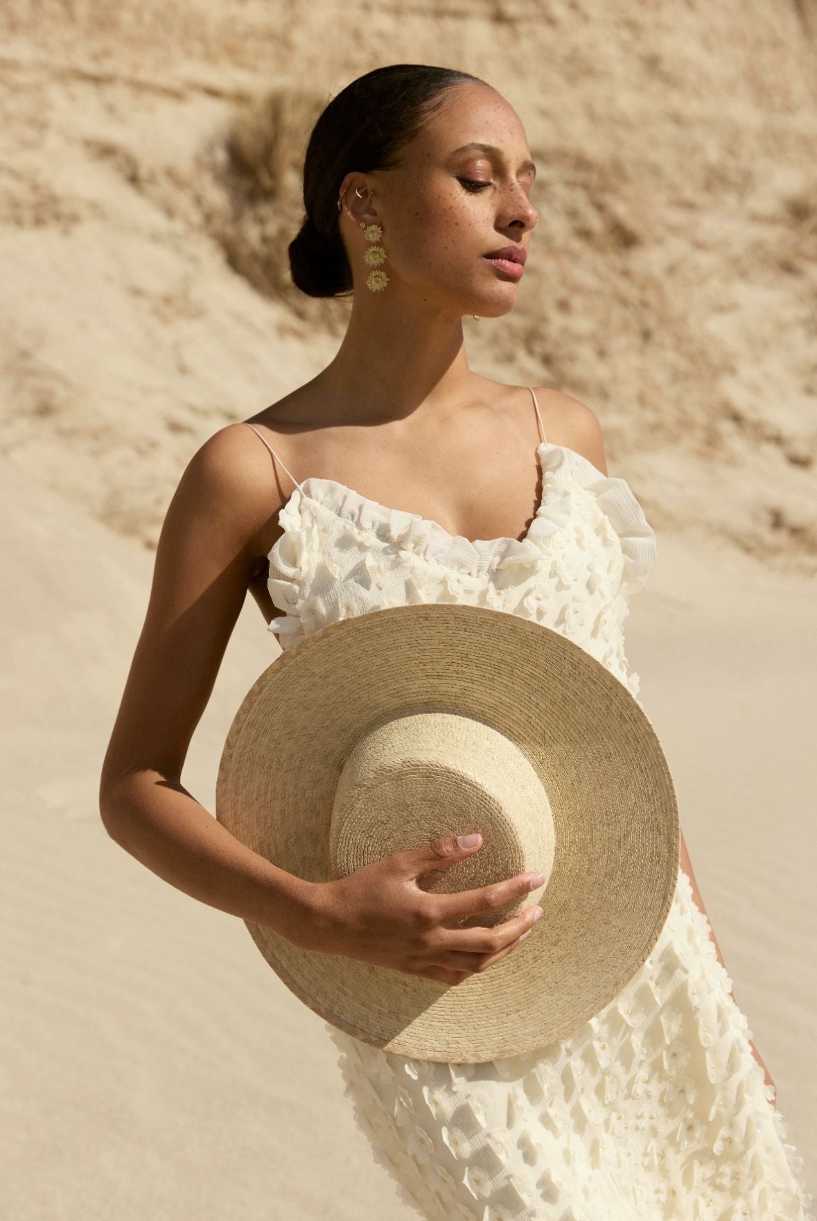 Femme en robe blanche et chapeau assise sur la plage.