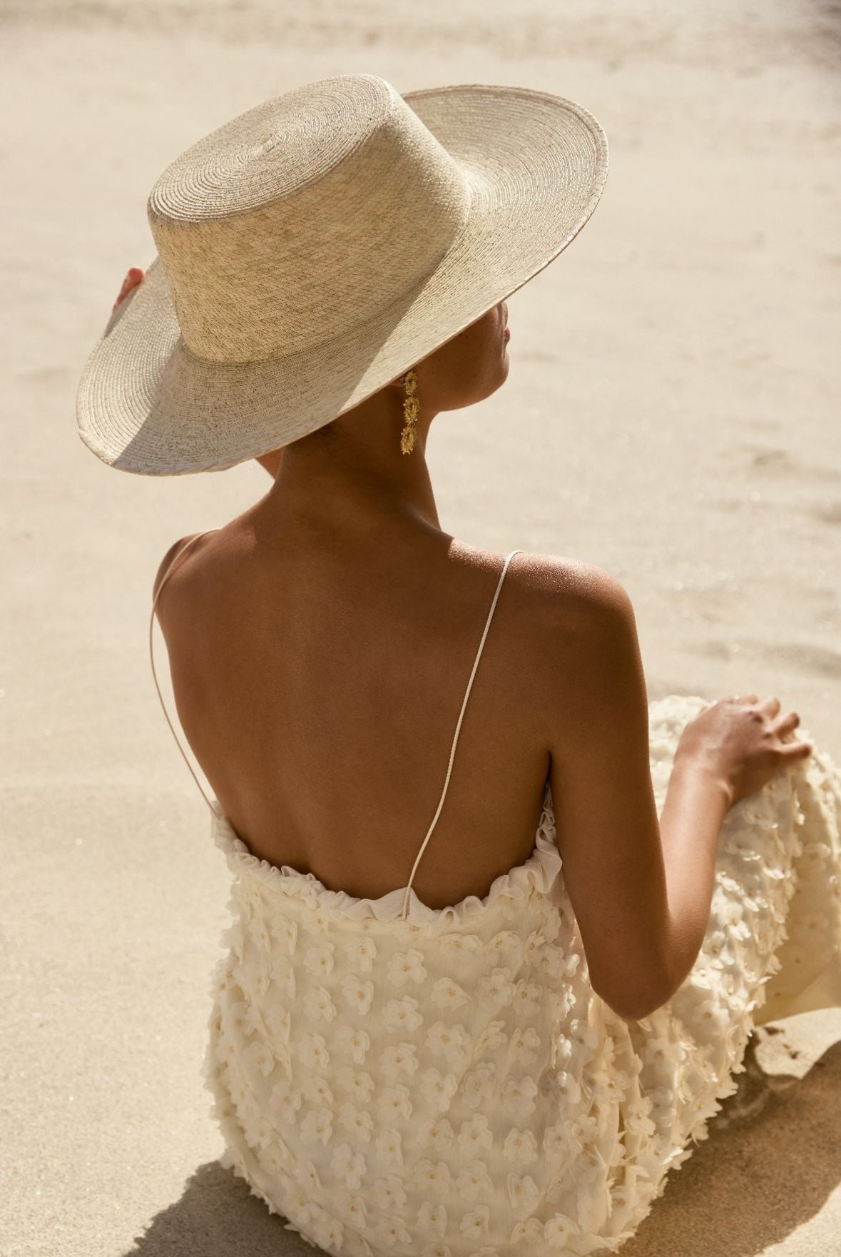 Femme de dos en robe blanche et chapeau assise sur la plage. 