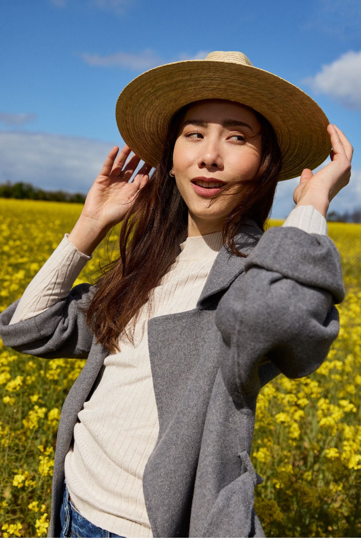 femme souriante en chapeau de paille et manteau se tenant dans un champ de fleurs jaunes.
