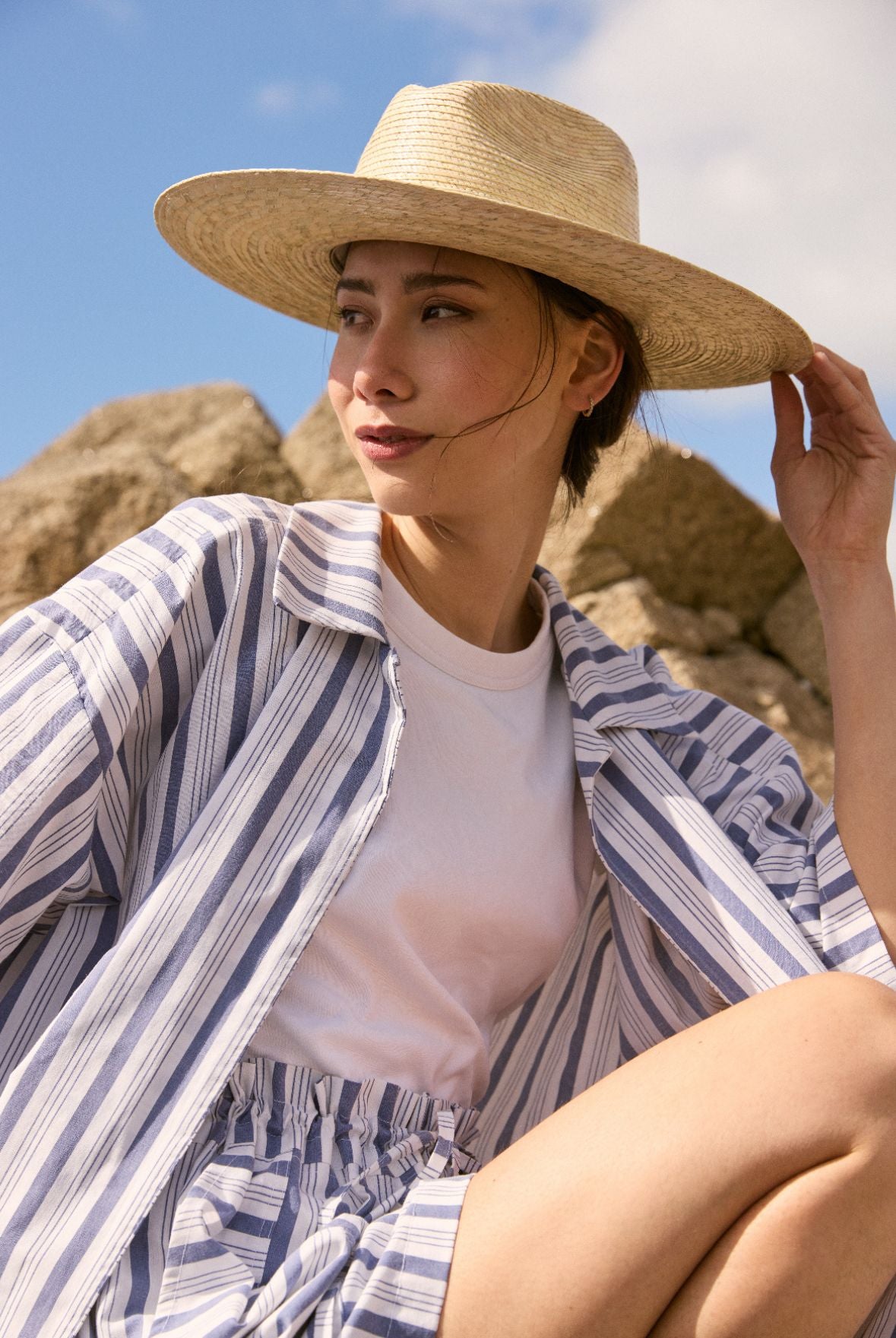 Une femme portant un chapeau de paille  et une chemise assise sur des rochers sous un ciel bleu