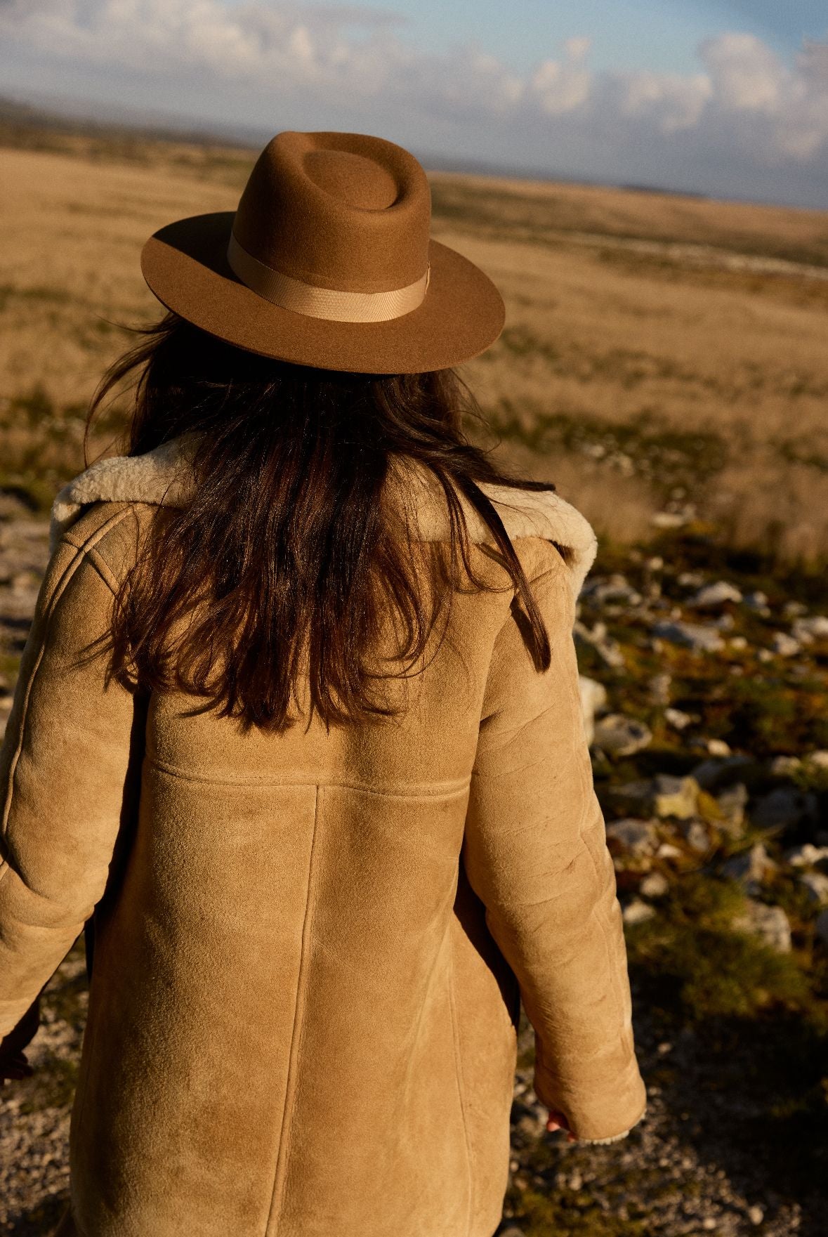 femme en manteau et chapeau bruns marchant sur une colline rocheuse
