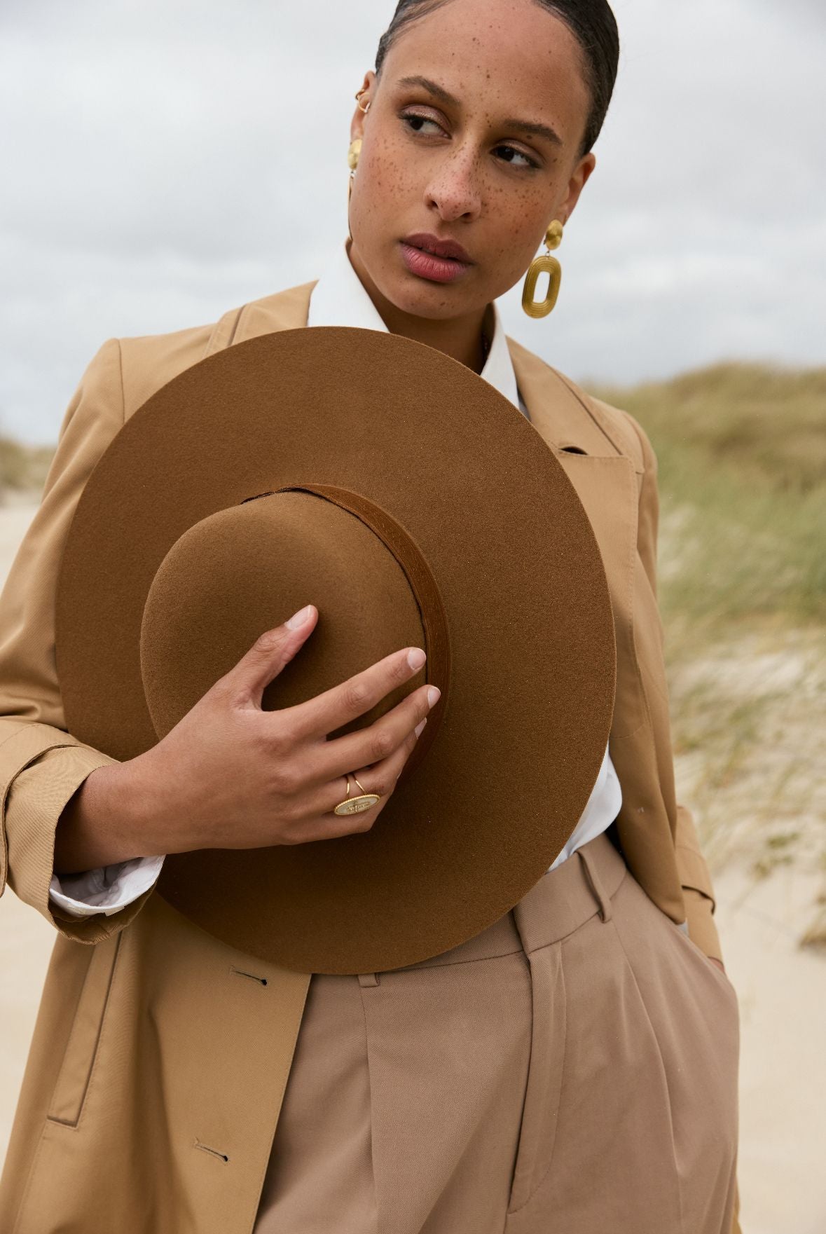 Une femme en manteau et chapeau marron debout dans le sable.
