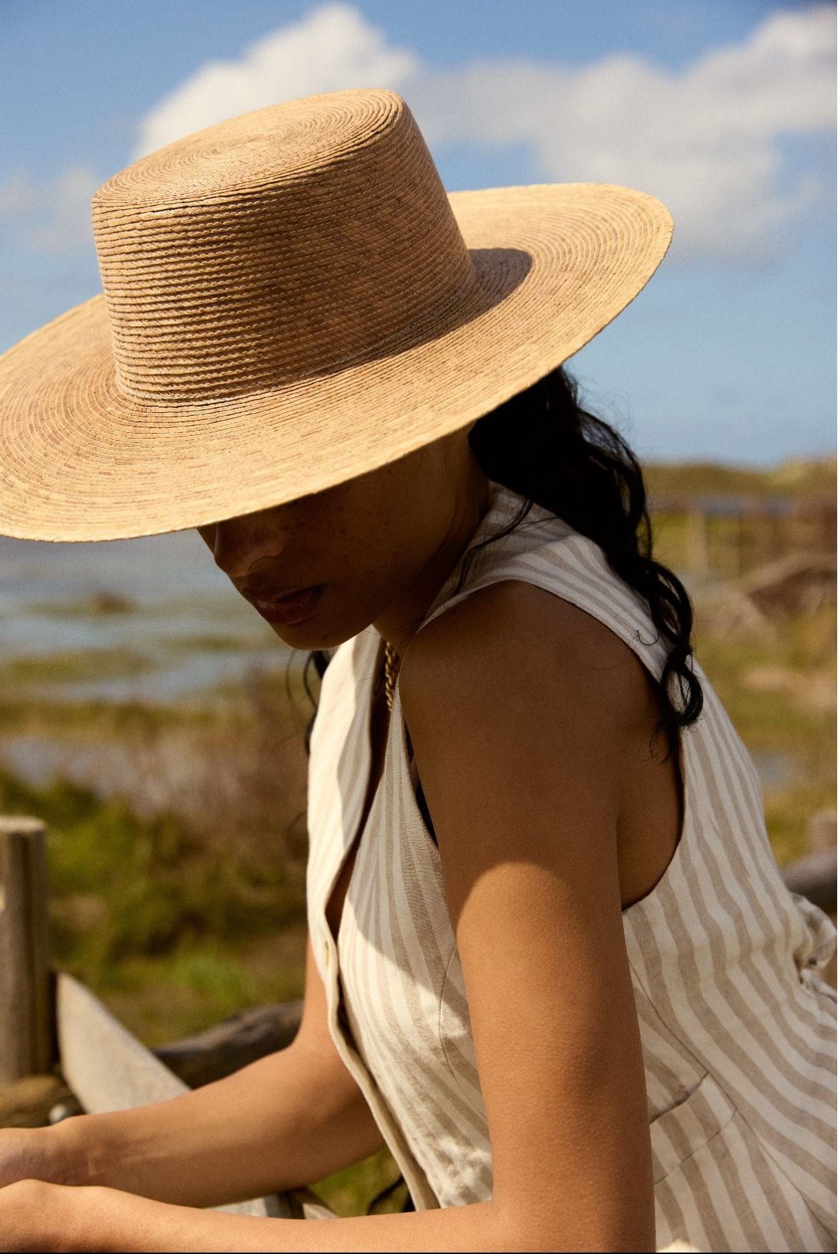 Femme en chapeau et chemise blanche appuyée contre une clôture sous un ciel bleu d'été.
